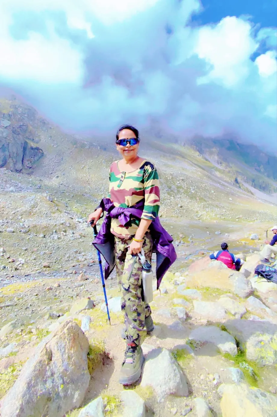 Roseleen smiling at the top of Hampta Pass with a trekking pole, rocky slopes and clouds behind her