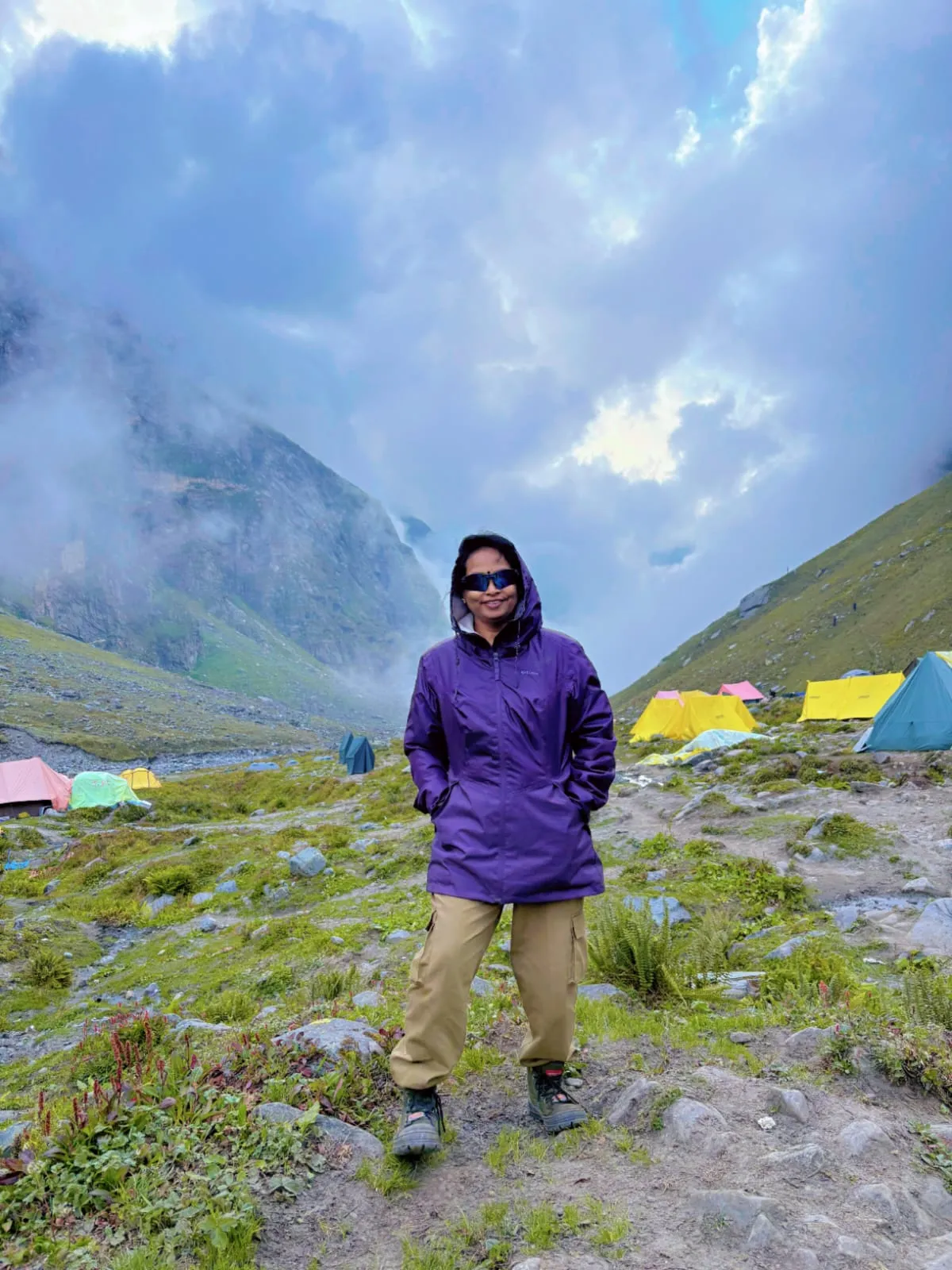 Roseleen in a purple rain jacket standing at the Balu Ka Ghera campsite with tents and cloud-covered mountains behind her