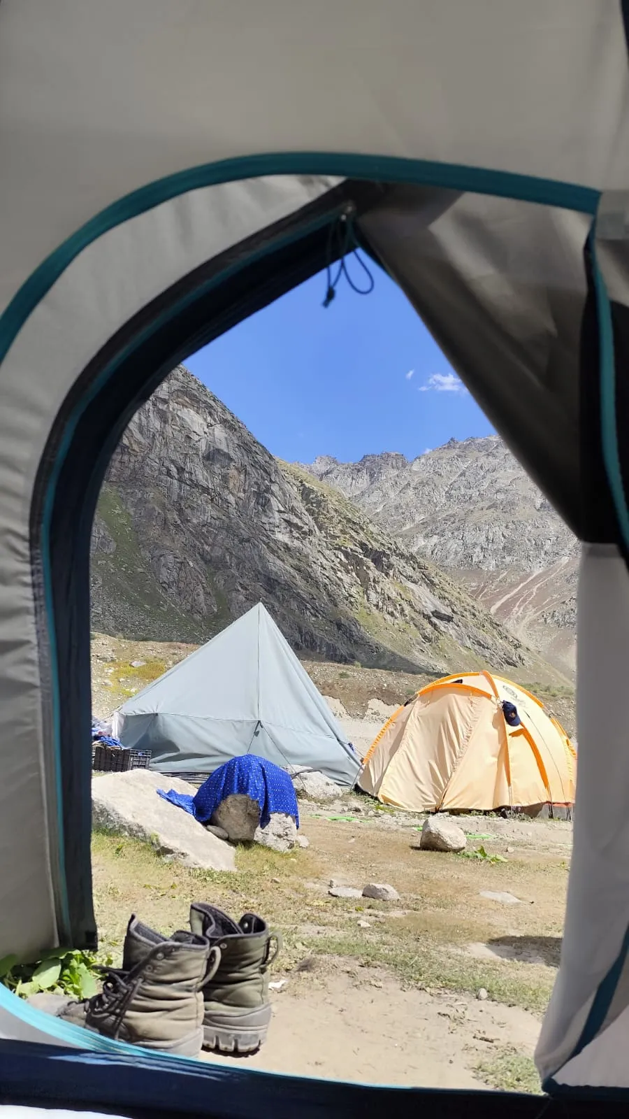 View from inside a tent at Chatru campsite showing trekking boots, camp tents and barren Lahaul mountains under a blue sky